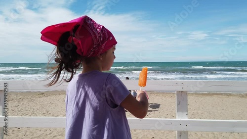 baby girl child eat popsicle at the beach bar on wind sea with pink bandana