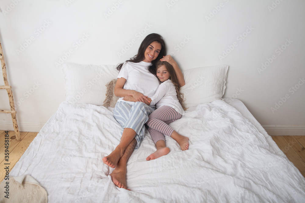 Happy loving family lying relaxing and enjoying in bedroom together. Mother and her cute preschool daughter child girl in pajamas playing and hugging in white bed in the morning. Happy mother's day. Happy loving family lying relaxing and enjoying in bedroom together. Mother and her cute preschool daughter child girl in pajamas playing and hugging in white bed in the morning. Happy mother's day.