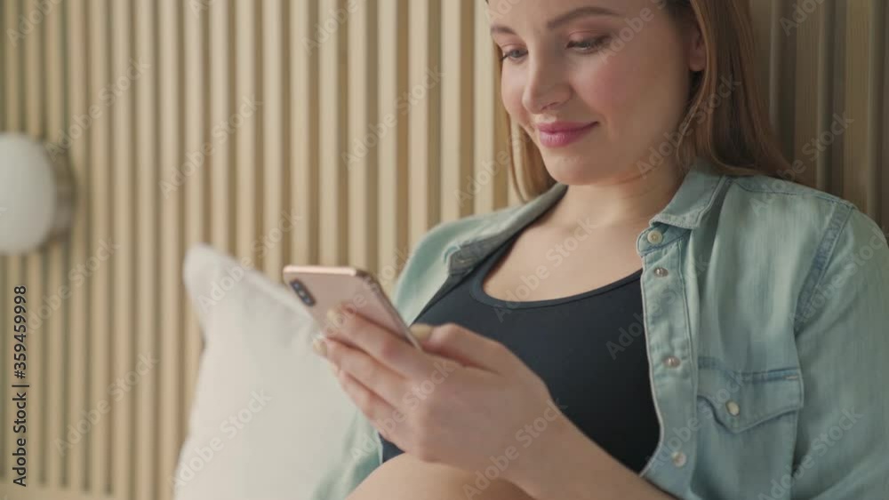 A close-up view of a beautiful pregnant woman is using her smartphone while lying on bed in sunny room A close-up view of a beautiful pregnant woman is using her smartphone while lying on bed in sunny room
