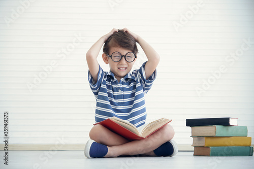 Obraz na plátně A little Caucasian boy wear eye glasses reading a book and sitting on the floor at home