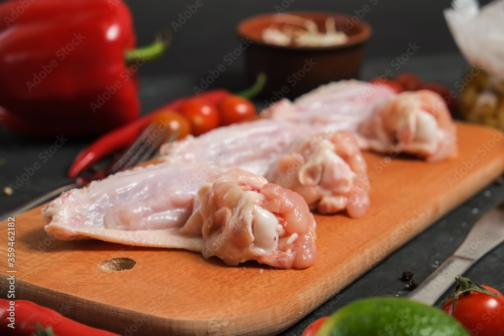Chicken wings in a wooden Cutting board, fresh rosemary, fresh cherry tomatoes, fresh chili peppers, garlic, lime and sea salt on a black background.