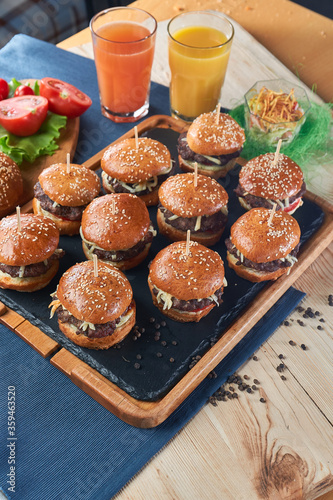 Juicy mini burgers with onion rings, jalapeno pepper, cucumbers, lettuce, cheese on a black stone background. Orange and carrot juce