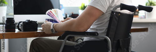 Close-up of professional designer working in office holding colourful palette. Male sitting in wheelchair. Photocamera and laptop on desktop. People with disability concept