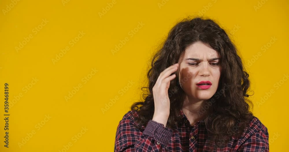 Close-up attractive plus-size model posing. A curly-haired brunette on bright yellow studio background demonstrating different emotions. Girl suffering from sudden headache and holds her head Close-up attractive plus-size model posing. A curly-haired brunette on bright yellow studio background demonstrating different emotions. Girl suffering from sudden headache and holds her head