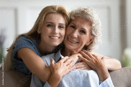 Different generation family relatives women portrait. Grandmother sit on sofa her grown up granddaughter hug her from behind express sincere feelings attachment protection love, close up concept photo