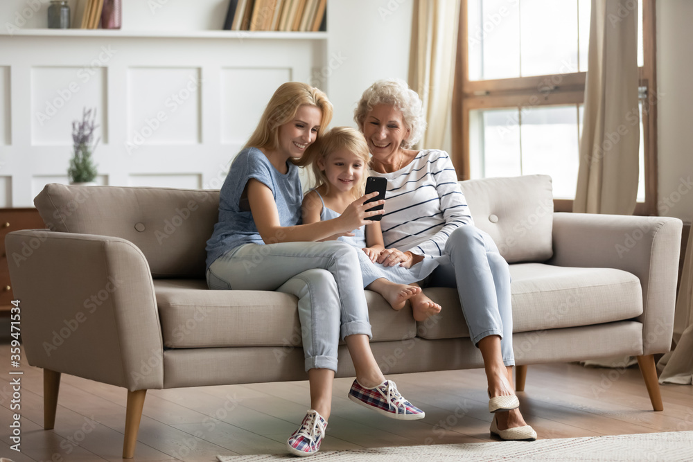 Three generations of women rest on couch have fun watch funny videos on  smartphone, happy little