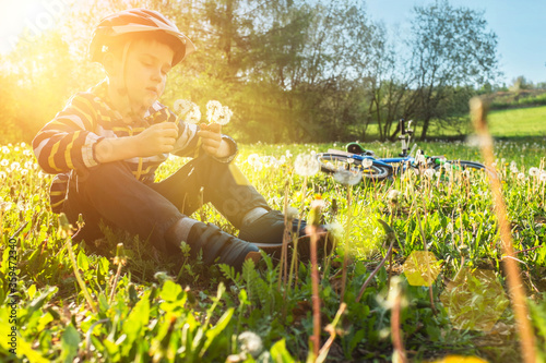 Child with a bouquet of dandelions sitting on grass in park. Boy going wearing safe bicycle helmets. Kid biking on sunny summer day. Active healthy outdoor sport. Fun activity.