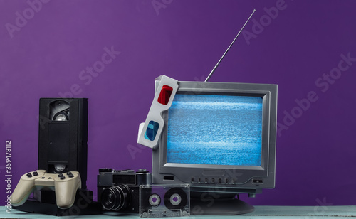 Retro media, entertainment 80s. Antenna old-fashioned retro tv receiver, anaglyph stereo glasses, audio and video cassette, gamepad, camera on purple background.