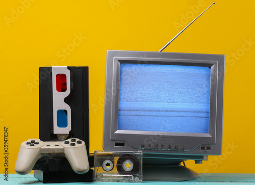 Retro media, entertainment 80s. Antenna old-fashioned retro tv receiver, anaglyph stereo glasses, audio and video cassette, gamepad on yellow background.