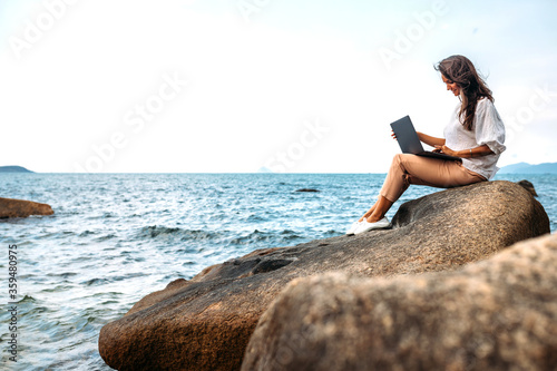 Young successful  smiling girl working using a laptop  sitting on a rocky seashore. traveling  blogger  freelancer  content plan  work online