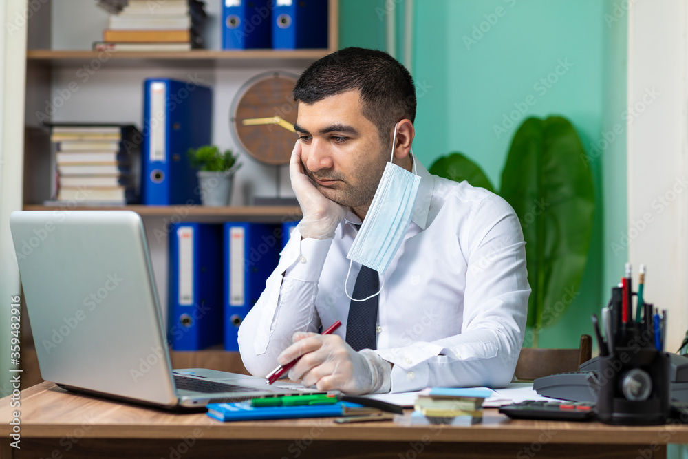 Tired young business man working on laptop with half worn medical protective mask and gloves