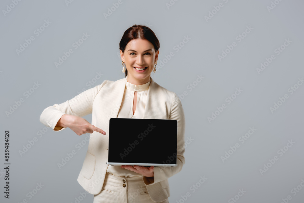 Beautiful businesswoman pointing with finger at laptop and smiling at camera isolated on grey