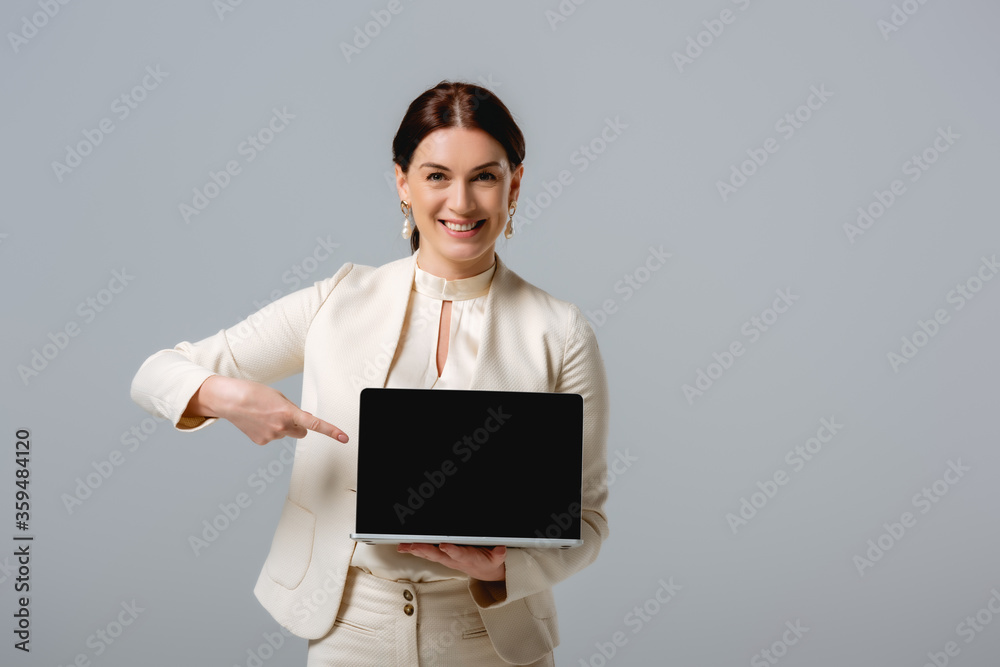 Beautiful businesswoman pointing with finger at laptop and smiling at camera isolated on grey Beautiful businesswoman pointing with finger at laptop and smiling at camera isolated on grey