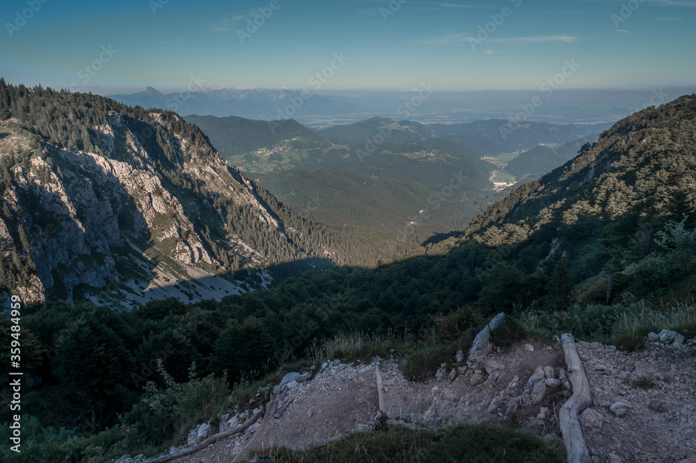 Fototapeta premium Aerial view of valley and Alps mountains, Slovenia. Hiking in summer season to the hilltop. Trail for hiking visible in foreground. Nature covered with green forest. Wide, elevated shot