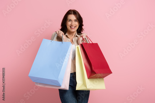Beautiful brunette woman smiling at camera while holding colorful shopping bags on pink background