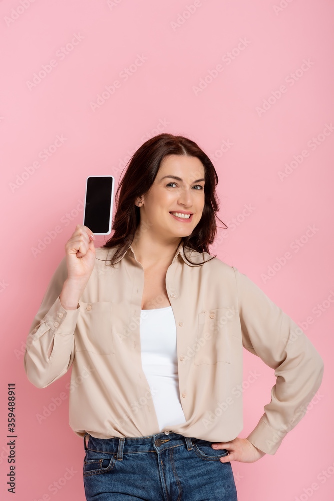 Beautiful smiling woman with hand on hip showing smartphone with blank screen isolated on pink