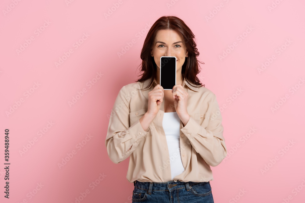 Obraz premium Brunette woman showing smartphone with blank screen near face on pink background