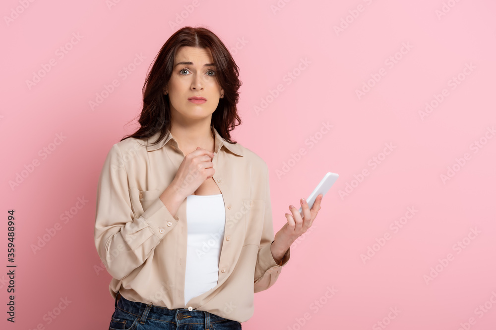 Worried woman looking at camera while holding smartphone on pink background Worried woman looking at camera while holding smartphone on pink background