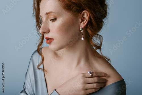 Fotografia Beautiful elegant redhead freckled woman wearing luxury  pearl jewelry: earrings, ring, silk blouse, posing in studio, on blue background