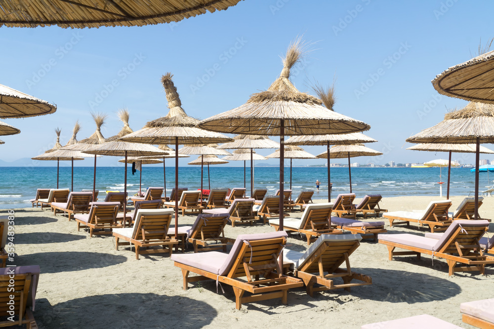 The rows of empty sunbeds with straw umbrellas on the beach. Blue sky and turquoise sea. Sunny summer day. Vlora / Vlore, Albania, Europe.