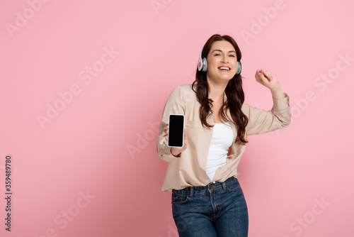 Cheerful woman in headphones showing smartphone on pink background