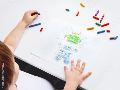 Left-handed toddler draws funny robot. Kid uses wax crayons. Top view on child's hands and pencils on white background. photo