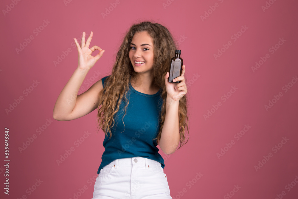 Happy girl with long curly hair, smiling holding bottle package of hair care product. Mockup with copy space, isolated on pink