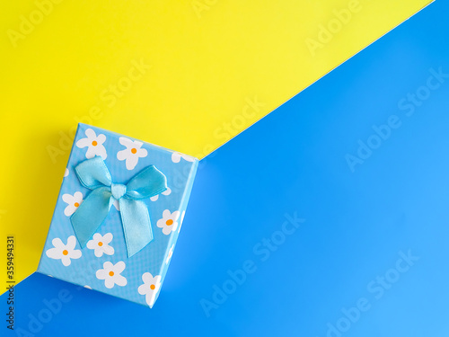 A light blue gift box tied with a gold ribbon. Top view on a contrasting background of bright blue and yellow paper lying diagonally. Place for text, postcards. Abstract background for design. © Ninaveter