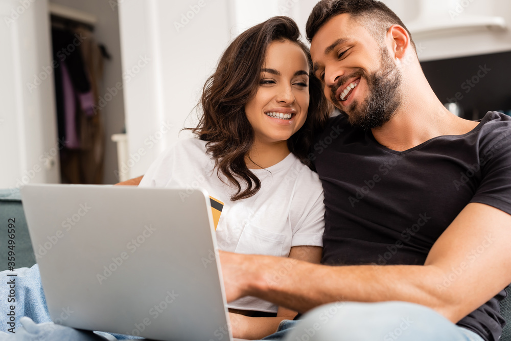 happy woman holding credit card near bearded boyfriend and laptop
