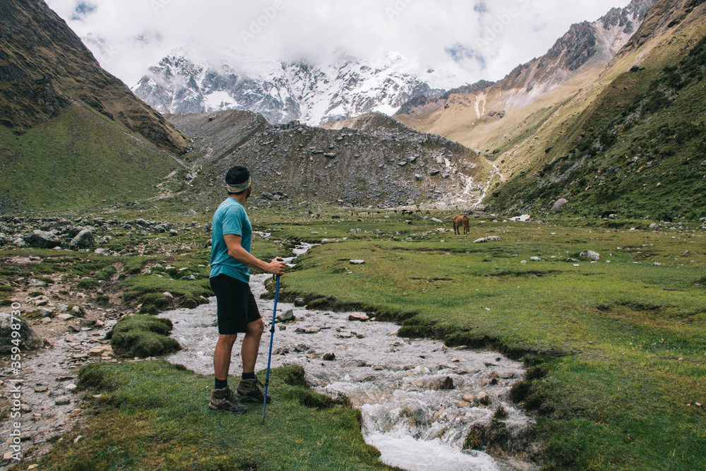 Back view of male traveler dressed in active wear holding trekking sticks and admiring wildness nature of high mountains.Experienced tourist enjoying wanderlust standing near river in Salkantay