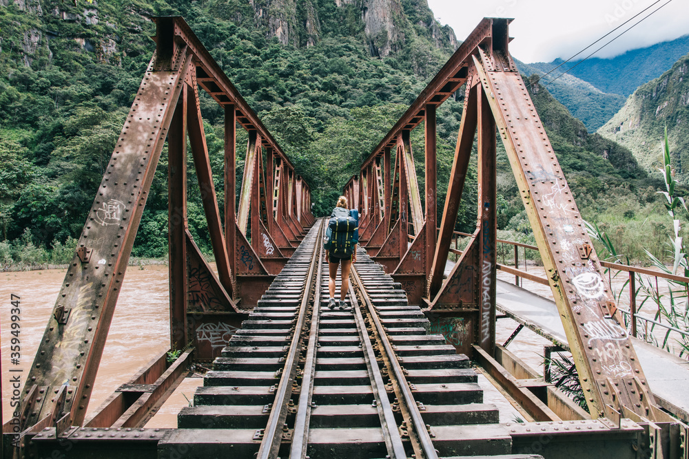 Back view of young woman tourist with backpack discover new places in wildness environment crossing bridge with rails over river.Hiker with travel rucksack going in mountains during trek