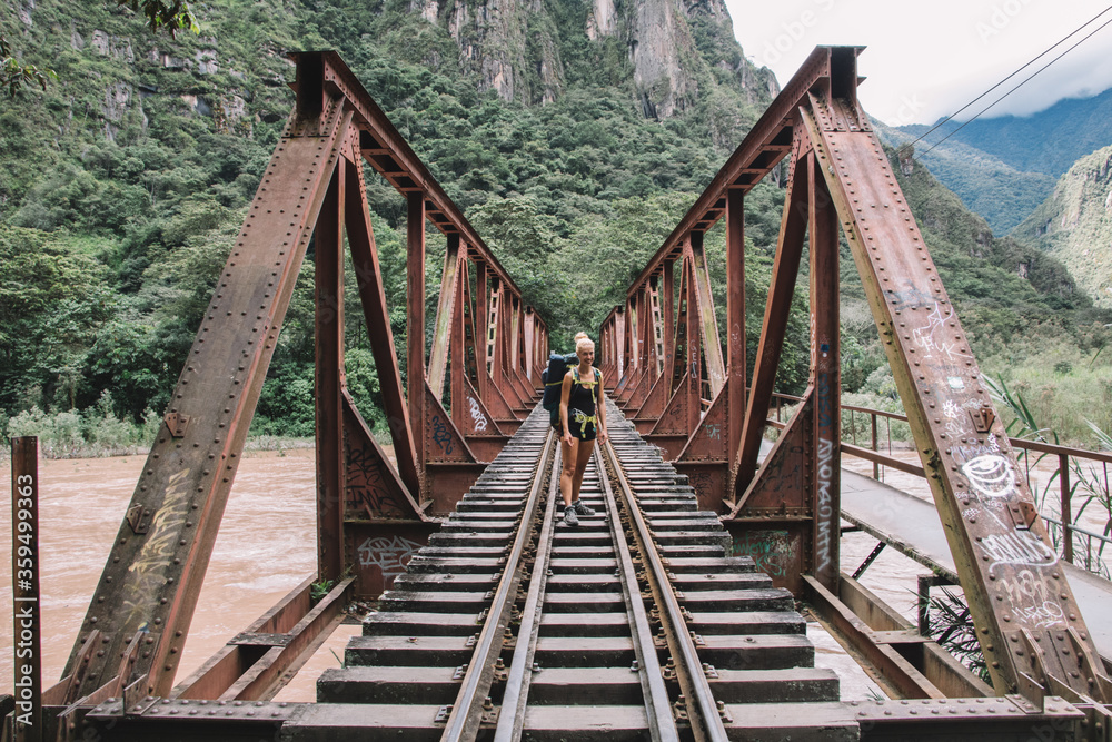 Portrait of positive young woman with tourist backpack smiling at camera and standing on rails of bridge while travelling in wildness nature with high mountains and beautiful scenery on background