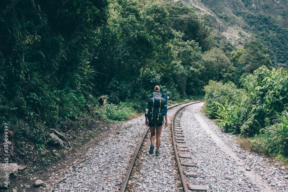 Fototapeta premium Rear view of female tourist with backpack discovering new places in wildness environment of mountains.Hiker with rucksack going on rainway and exploring green vegetation during trekking holiday