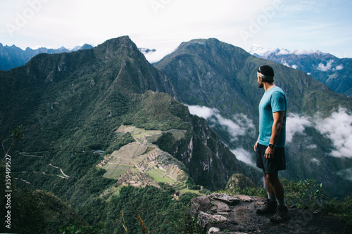 Professional young man dressed in active wear standing on peak of mountain and admiring green hills of Machu Picchu and breathtaking scenery of natural environment during trekking wanderlust