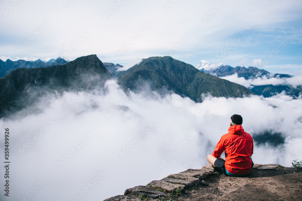 Obraz premium Back view of young man dressed in active wear sitting in lotus on edge of the summit enjoying breathtaking view of high green mountains covered white fog during trekking trip on holiday