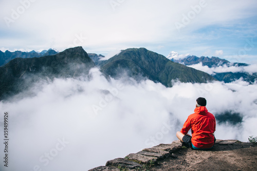 Back view of young man dressed in active wear sitting in lotus on edge of the summit enjoying breathtaking view of high green mountains covered white fog during trekking trip on holiday