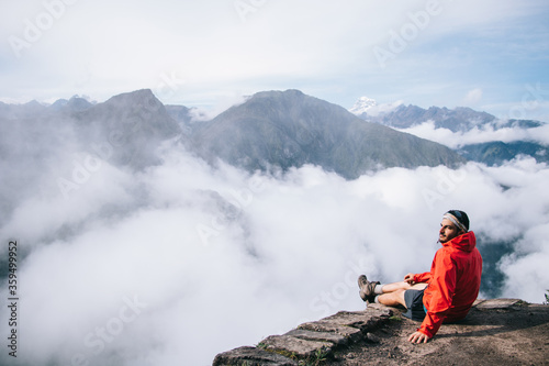 Experienced young man dressed in active wear relaxing on edge of summit enjoying calm of mountains and amazing view of natural environment during trip.Male tourist admiring breathtaking scenery