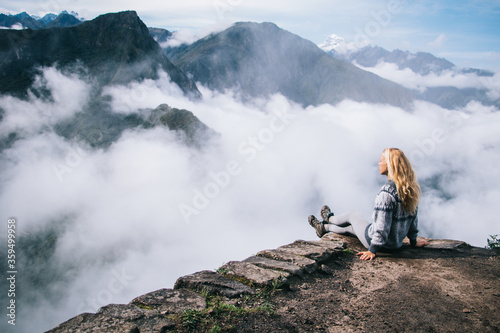 Fototapeta Naklejka Na Ścianę i Meble -  Blonde young woman traveler enjoying calm during trek sitting on edge of summit and thinking about inspiration.Female tourist relaxing and admiring breathtaking view of green mountains covered fog