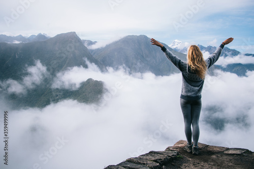 Back view of young woman tourist holding hands up enjoying freedom and travel lifestyle.Female hiker admiring beautiful scenery of green high mountains covered white mist standing on edge of summit