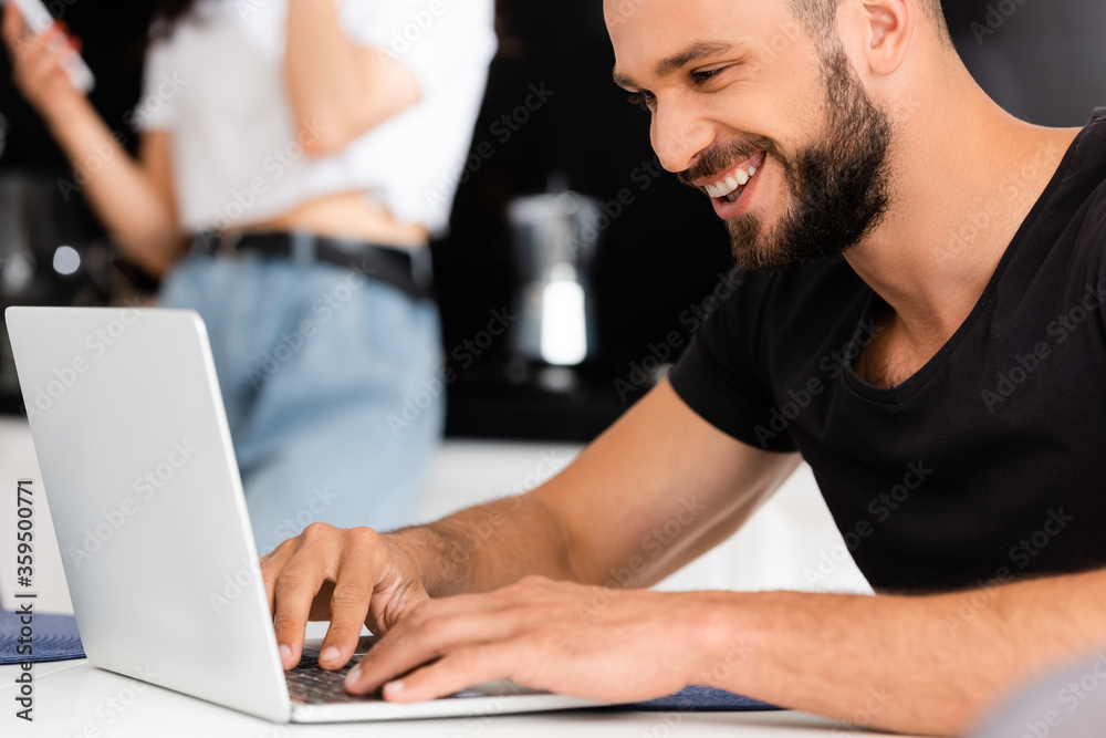 selective focus of cheerful freelancer using laptop near girlfriend in kitchen selective focus of cheerful freelancer using laptop near girlfriend in kitchen