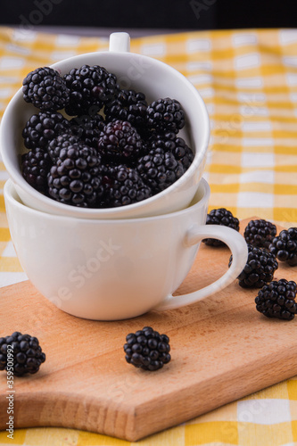 White cup with blackberries. Scattered berries on wooden kitchen board.