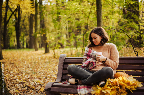 A girl holds a coffee and sits on a bench in the park in the fall. on the bench beautiful autumn yellow leaves and pumpkin