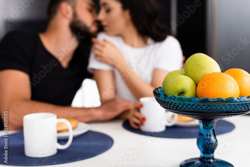selective focus of ripe fruits and cups near couple kissing in kitchen
