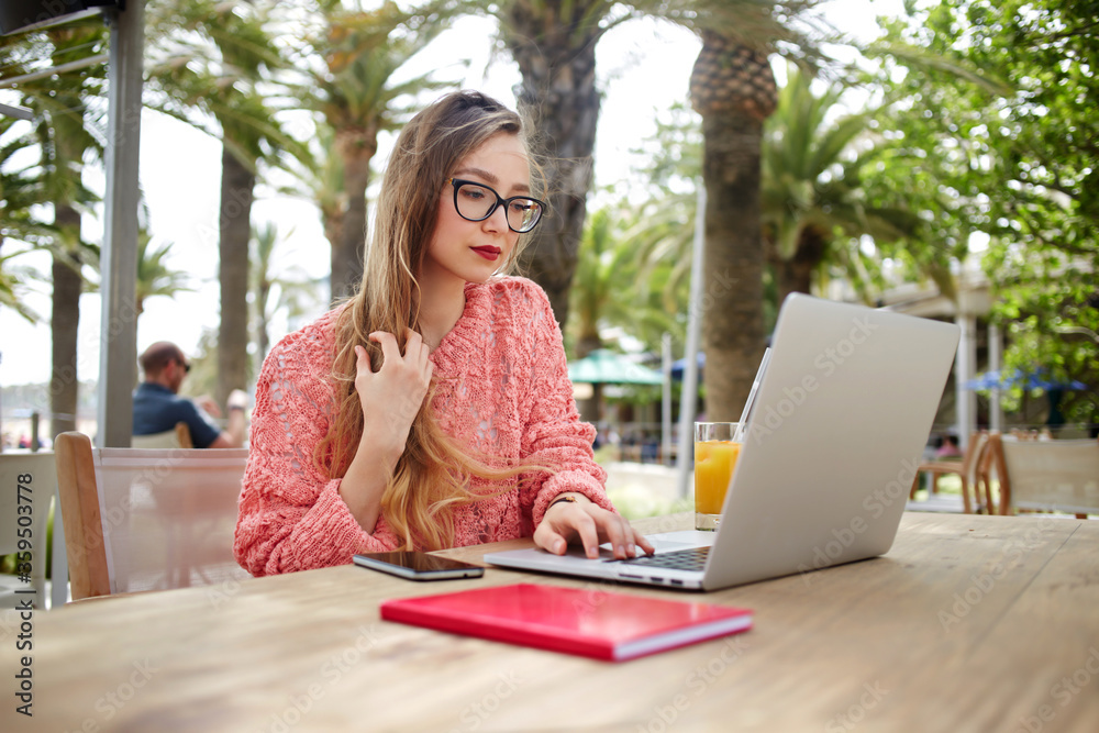 Portrait of young female freelancer working on laptop computer while sitting in sidewalk cafe against beautiful palm trees, smart women using net-book during morning breakfast in coffee shop outdoors Portrait of young female freelancer working on laptop computer while sitting in sidewalk cafe against beautiful palm trees, smart women using net-book during morning breakfast in coffee shop outdoors