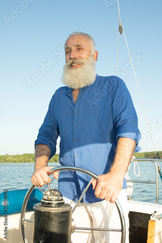 Mature bearded man standing at helm of yacht out at lake, steering, smiling.