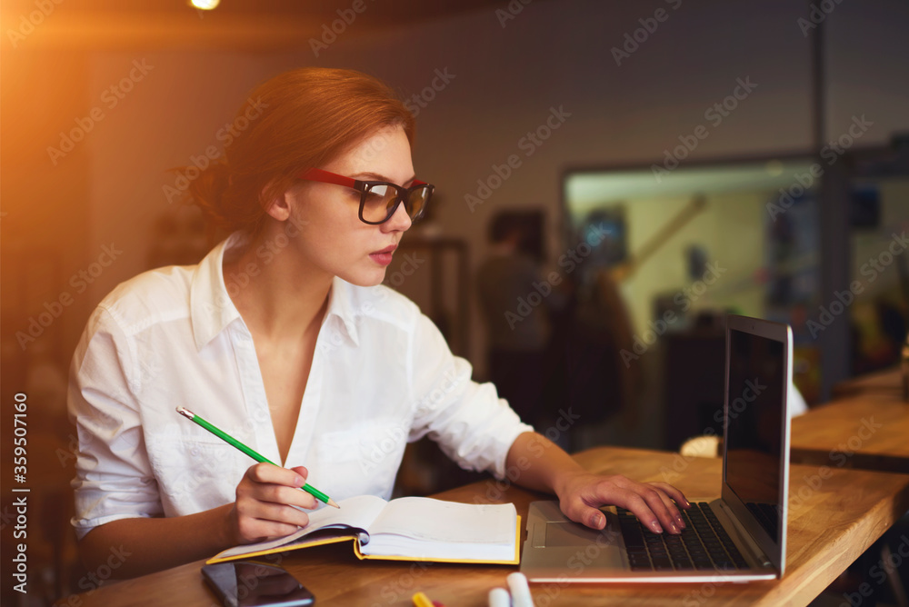 Skilled beautiful female graphic designer in spectacles making research on websites via laptop connected to wireless internet and writing useful information to textbook during free time in cafeteria Skilled beautiful female graphic designer in spectacles making research on websites via laptop connected to wireless internet and writing useful information to textbook during free time in cafeteria