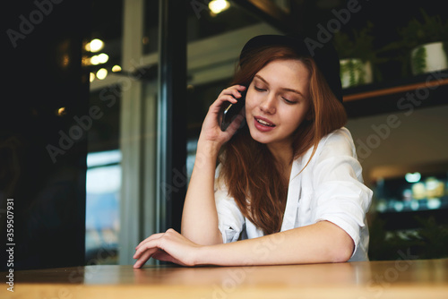 Charming young hipster girl in trendy hat having phone conversation with friend while sitting in cafe interior, positive female student contact banking service for checking money transfer on leisure