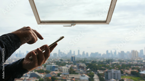 A man types a message on a smartphone against the background of an open window and a panorama of the city of Jakarta. Hands close up.