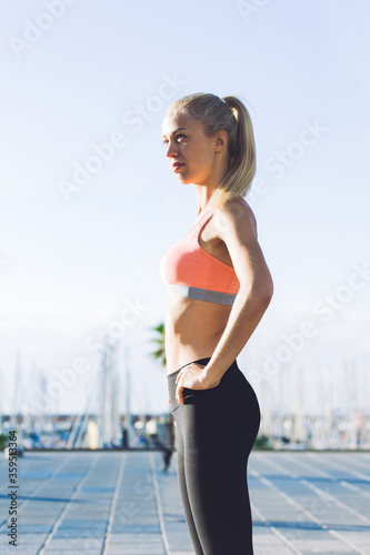 Half length portrait of a young female runner with perfect slender body taking break after workout outdoors, charming sports woman dressed in sportswear rest after active physical activity outside