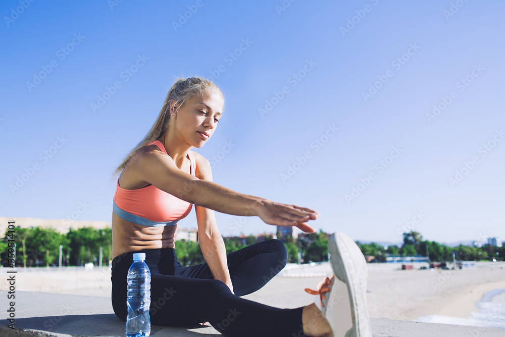 Young fit woman stretching leg while sitting on the concrete pier against blue sky background with copy space area for your text message or advertising content, female jogger working out outdoors